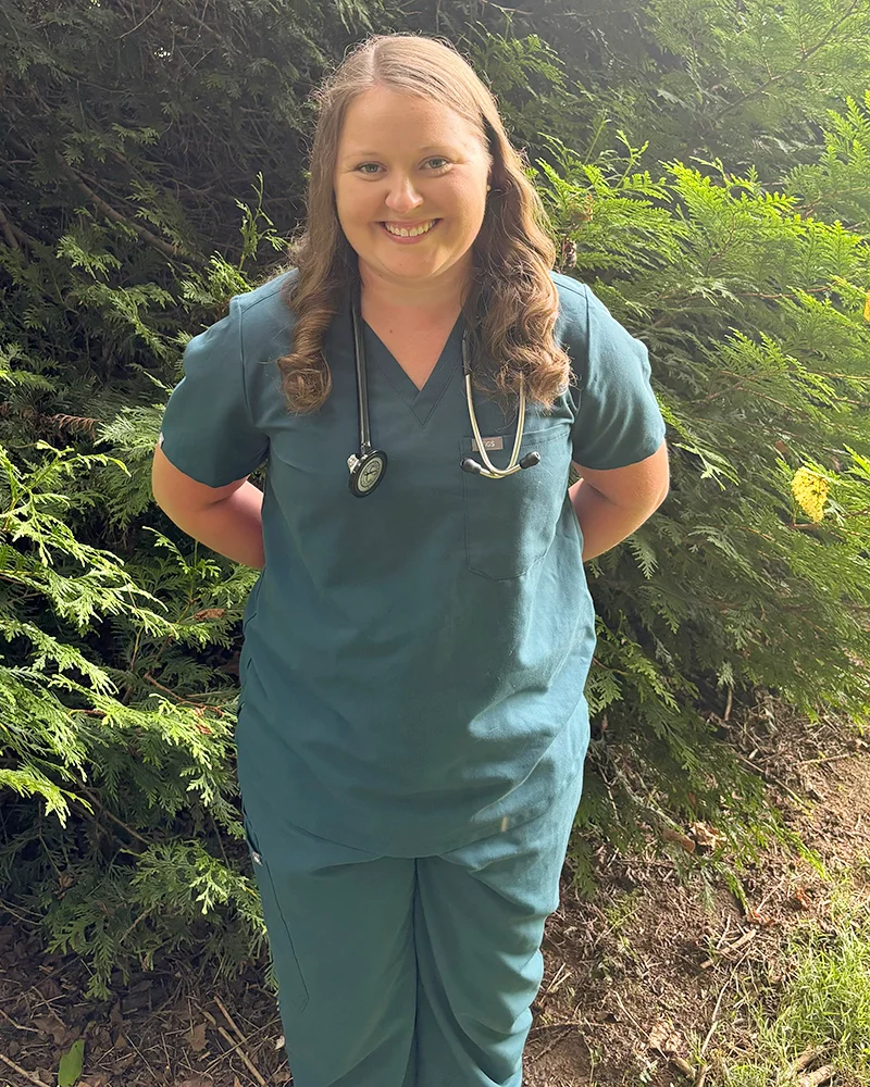 Dr. Anna Richardson, a veterinarian, is standing outdoors in front of lush green foliage. She is wearing teal scrubs with a stethoscope draped around her neck and smiling warmly at the camera. The natural setting and her relaxed posture give the impression of a caring and approachable professional.