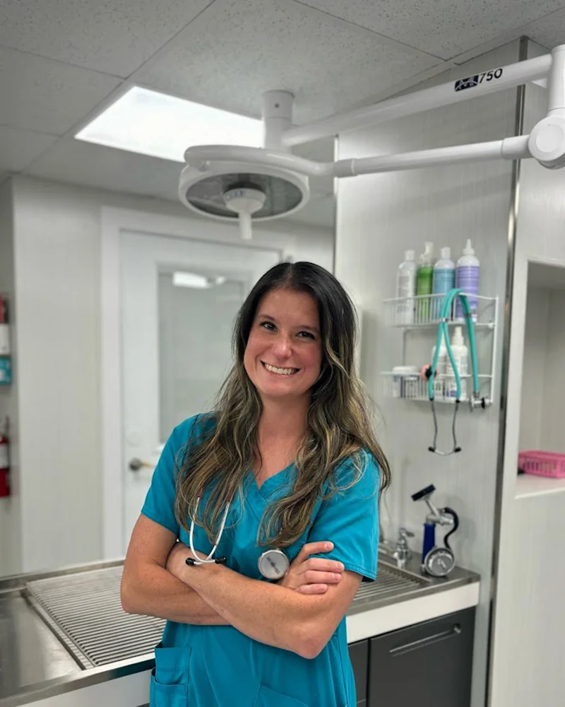 Dr. Danielle Reece, a veterinarian, is standing confidently with her arms crossed in a veterinary clinic. She is wearing teal scrubs with a stethoscope around her neck and smiling warmly at the camera. The background features medical equipment and supplies, indicating a professional and clean clinical setting.